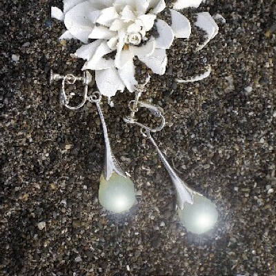 植物のつぼみをイメージした、緑色の石がセットされたシルバーピアス。白い花の模型と一緒に砂浜に置かれている。